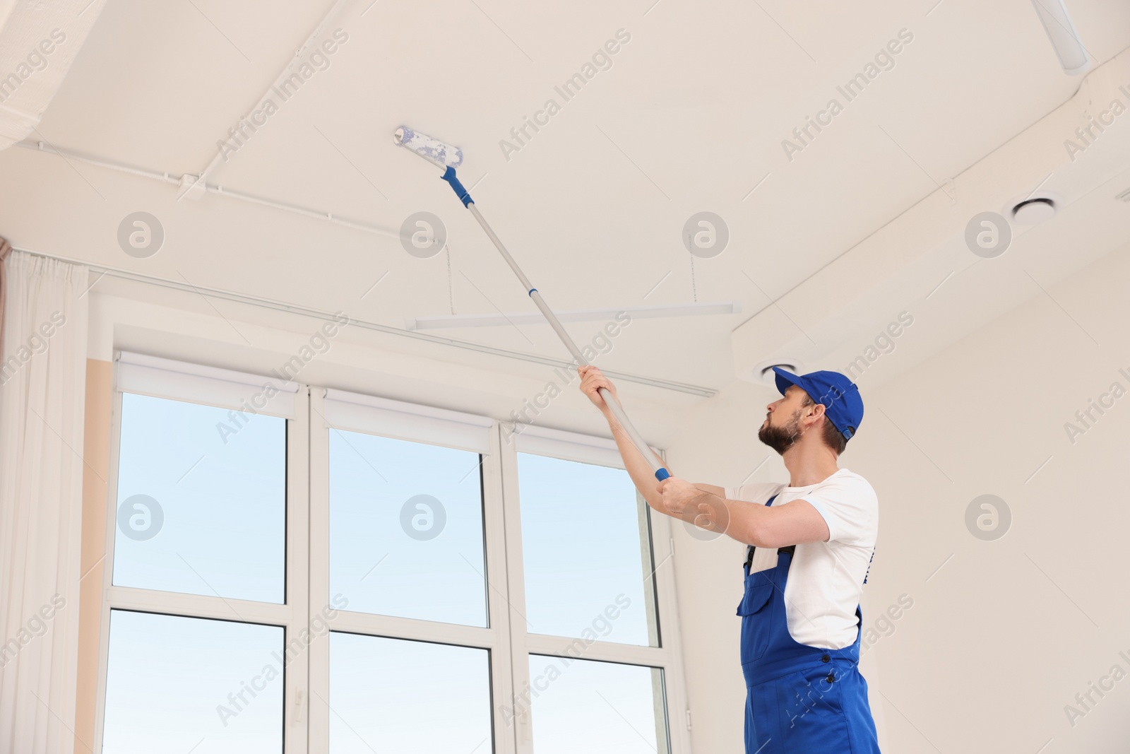 Photo of Worker in uniform painting ceiling with roller indoors, low angle view