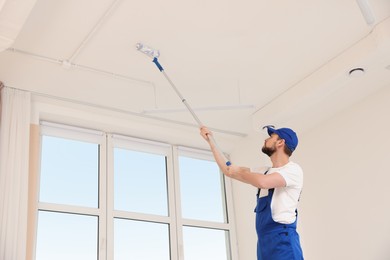 Photo of Worker in uniform painting ceiling with roller indoors, low angle view