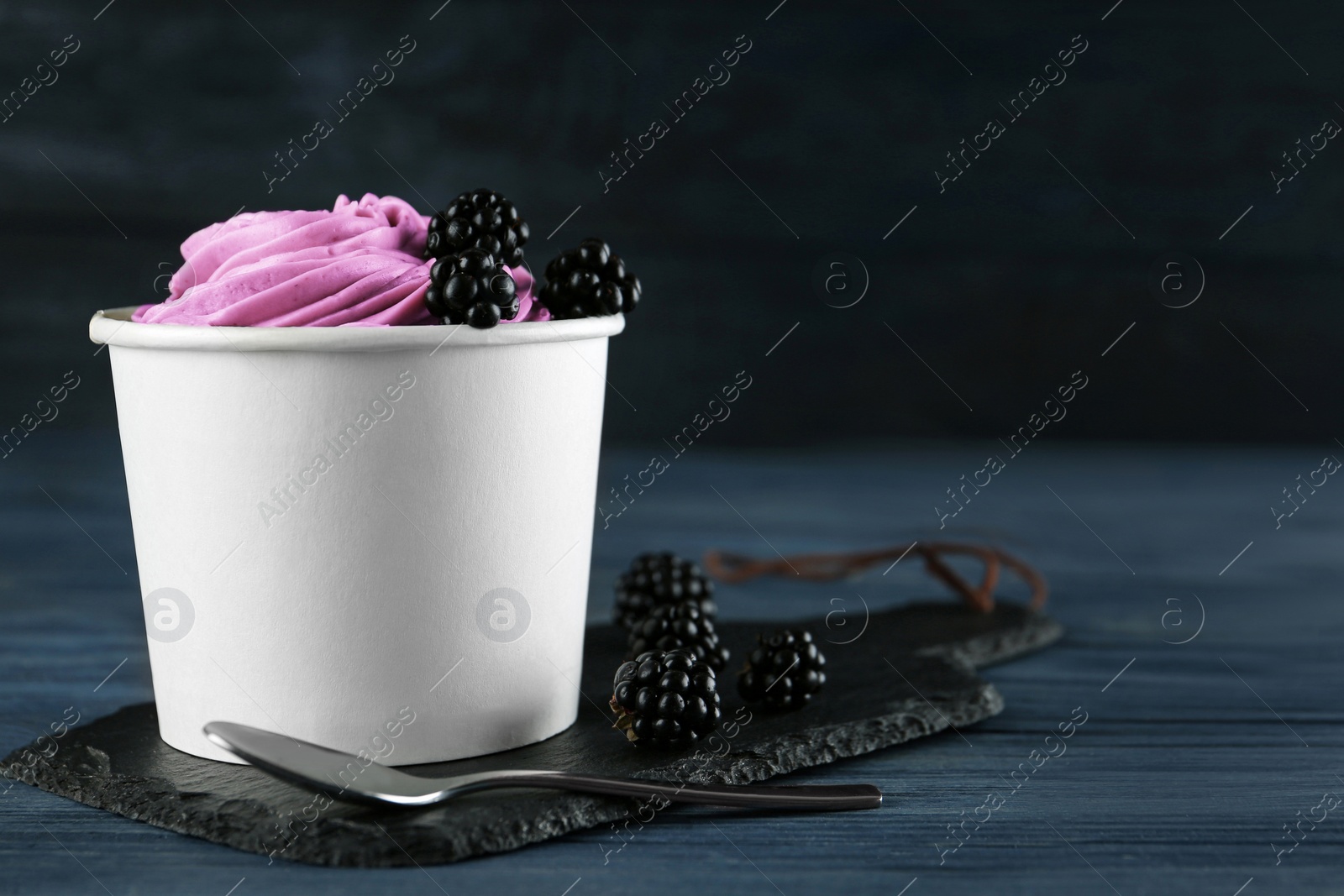 Photo of Cup with tasty frozen yogurt and blackberries on blue wooden table. Space for text