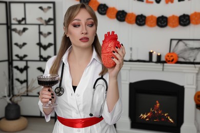 Woman in scary nurse costume with glass of wine and heart model indoors, space for text. Halloween celebration