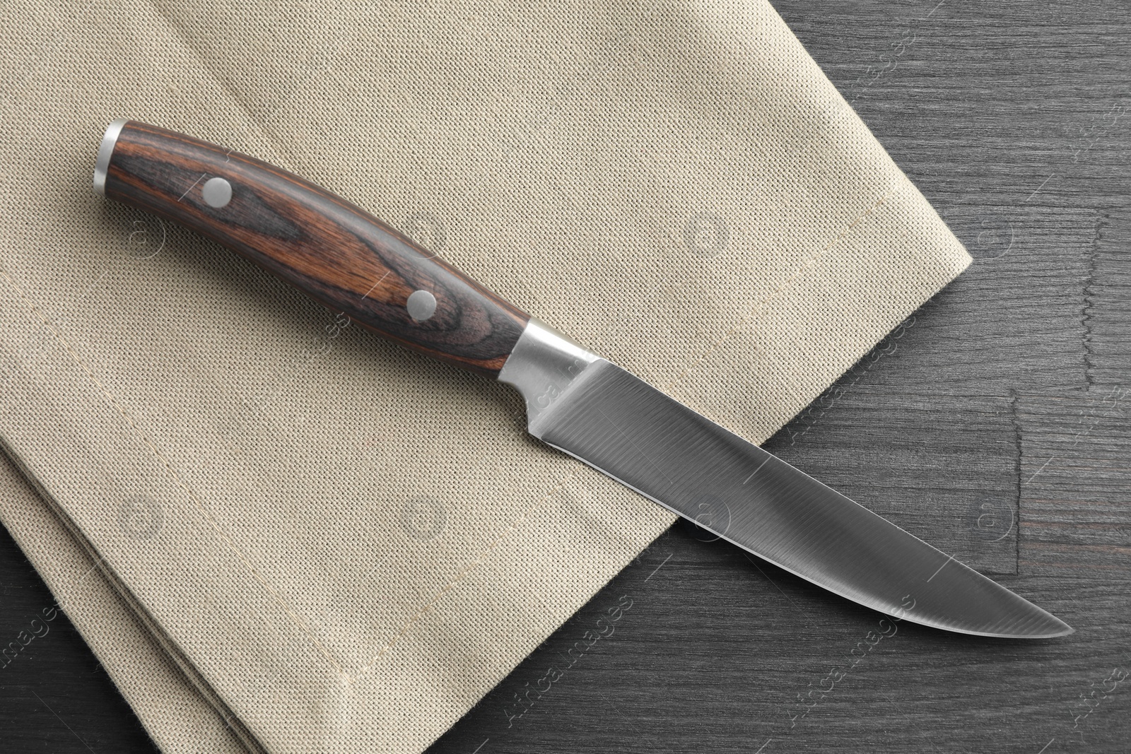 Photo of One sharp knife and napkin on black wooden table, top view