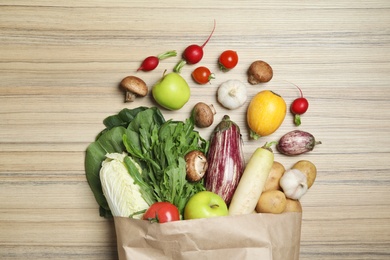 Overturned paper bag with vegetables and apples on wooden background, flat lay