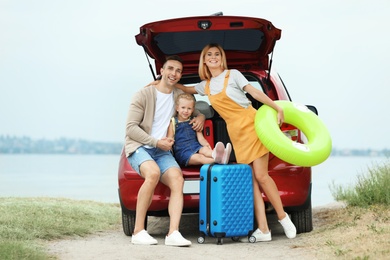 Happy family with suitcases and inflatable ring near car on riverside