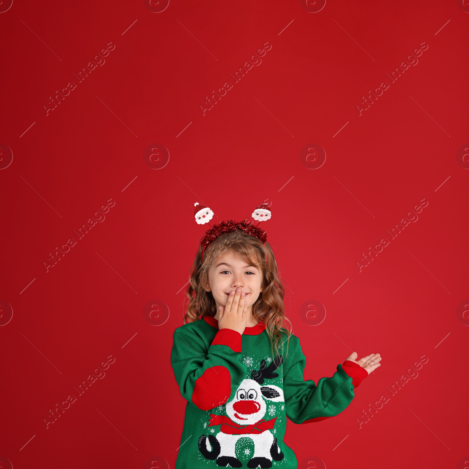 Photo of Cute little girl in green Christmas sweater covering her mouth against red background