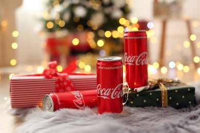 Photo of MYKOLAIV, UKRAINE - January 01, 2021: Cans of Coca-Cola and gifts on faux fur against blurred Christmas lights