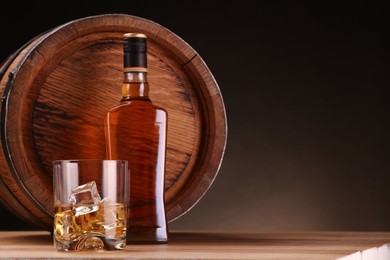 Photo of Whiskey with ice cubes in glass, bottle and barrel on wooden table against dark background, space for text