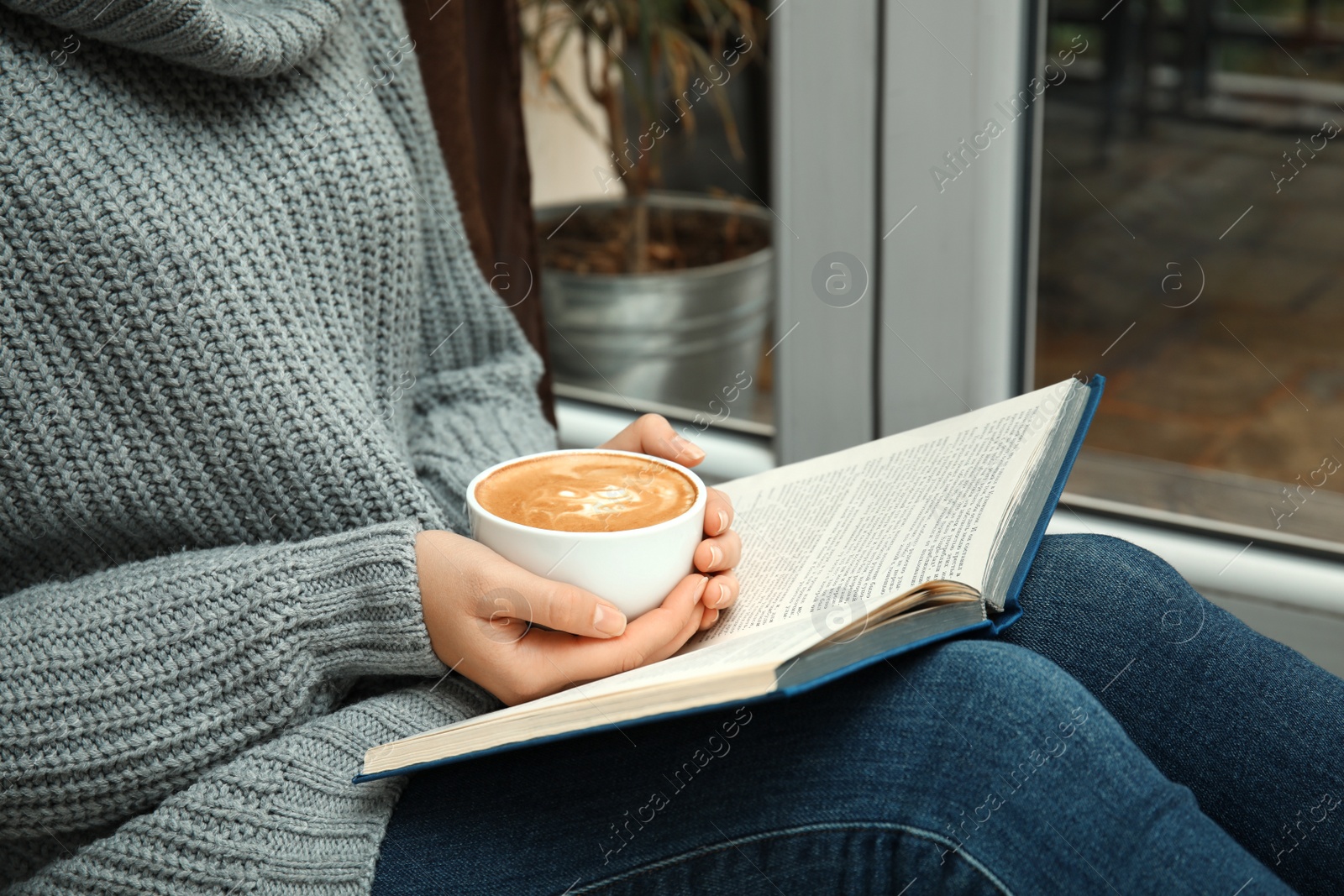 Photo of Woman with cup of coffee reading book near window indoors, closeup