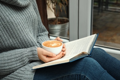 Photo of Woman with cup of coffee reading book near window indoors, closeup