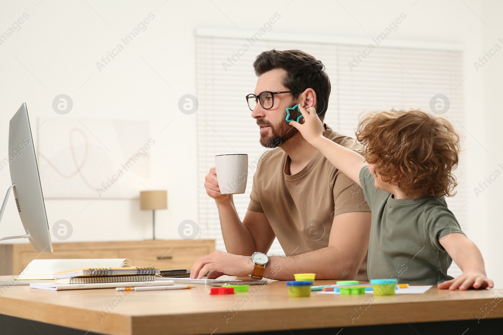 Photo of Little boy bothering his father at home. Man working remotely at desk