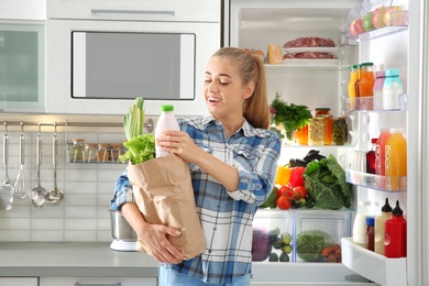 Woman with paper bag full of products standing near refrigerator in kitchen