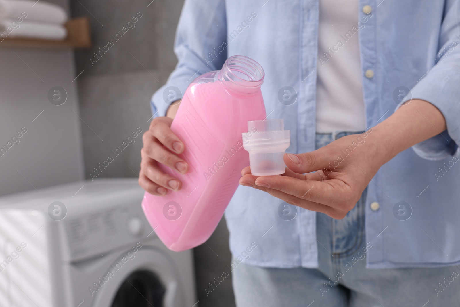 Photo of Woman pouring fabric softener from bottle into cap for washing clothes indoors, closeup