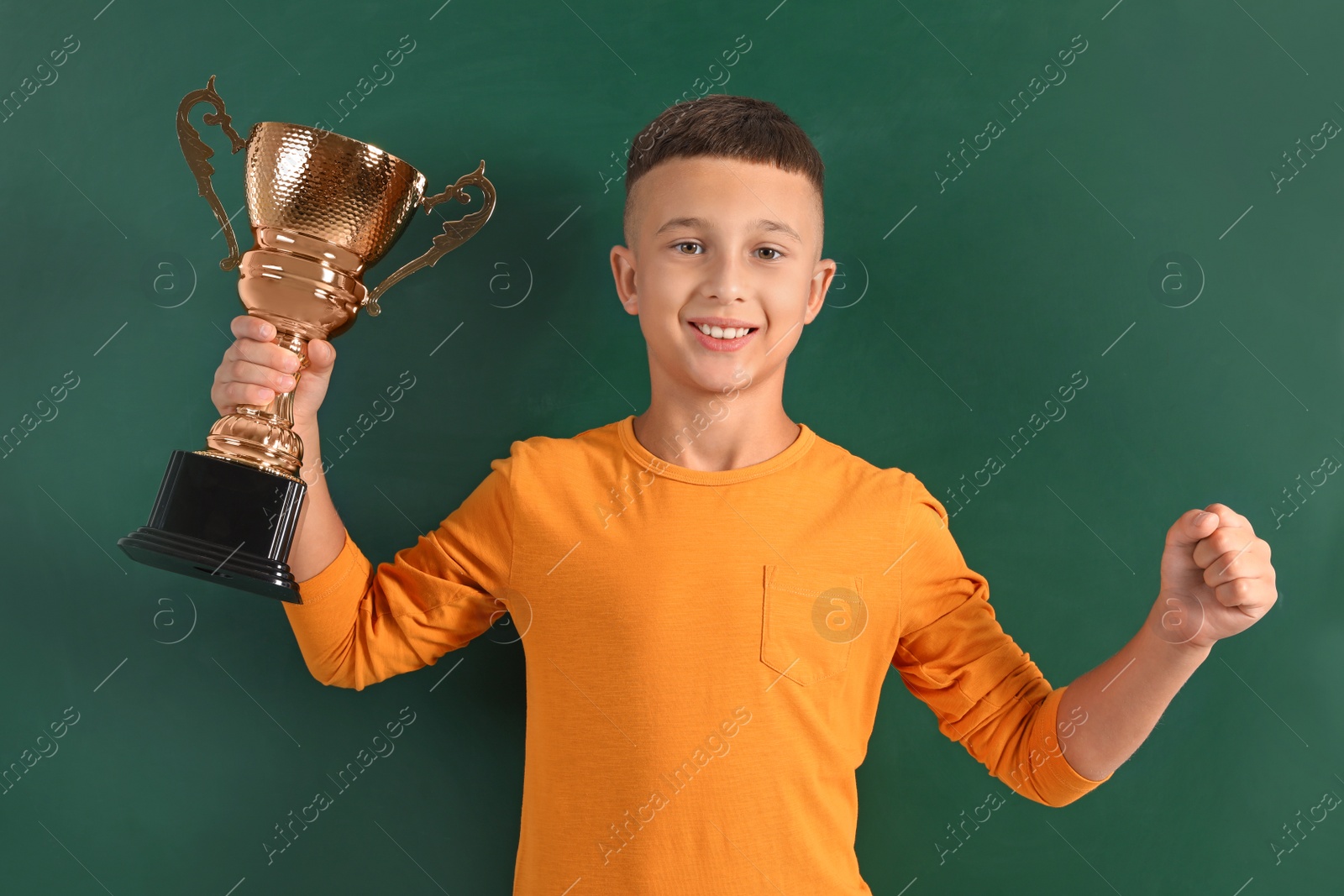 Photo of Happy boy with golden winning cup on near chalkboard