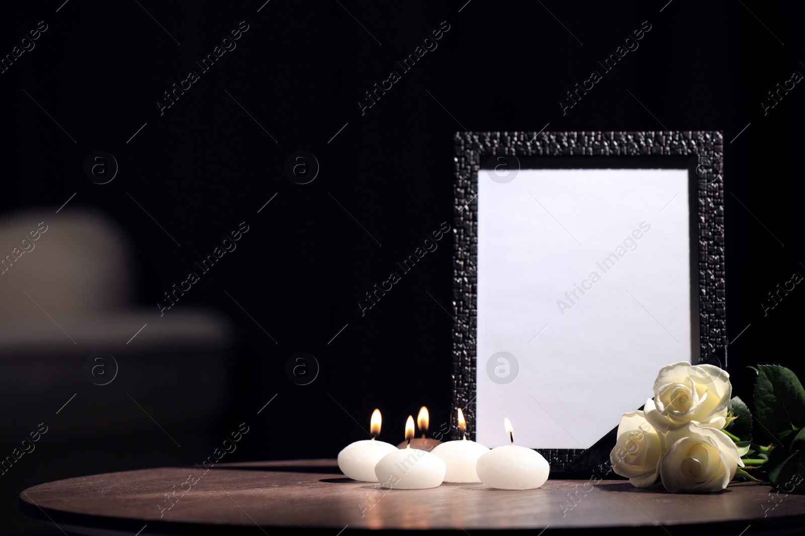Photo of Funeral photo frame with black ribbon, roses and candles on wooden table indoors. Space for design