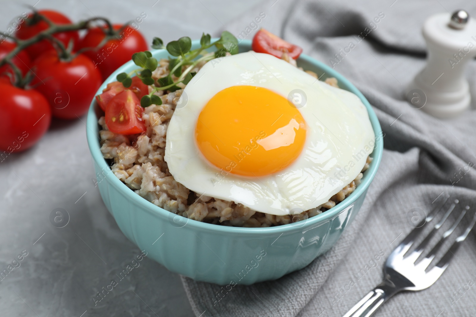 Photo of Tasty boiled oatmeal with fried egg, tomato and microgreens served on light grey table, closeup
