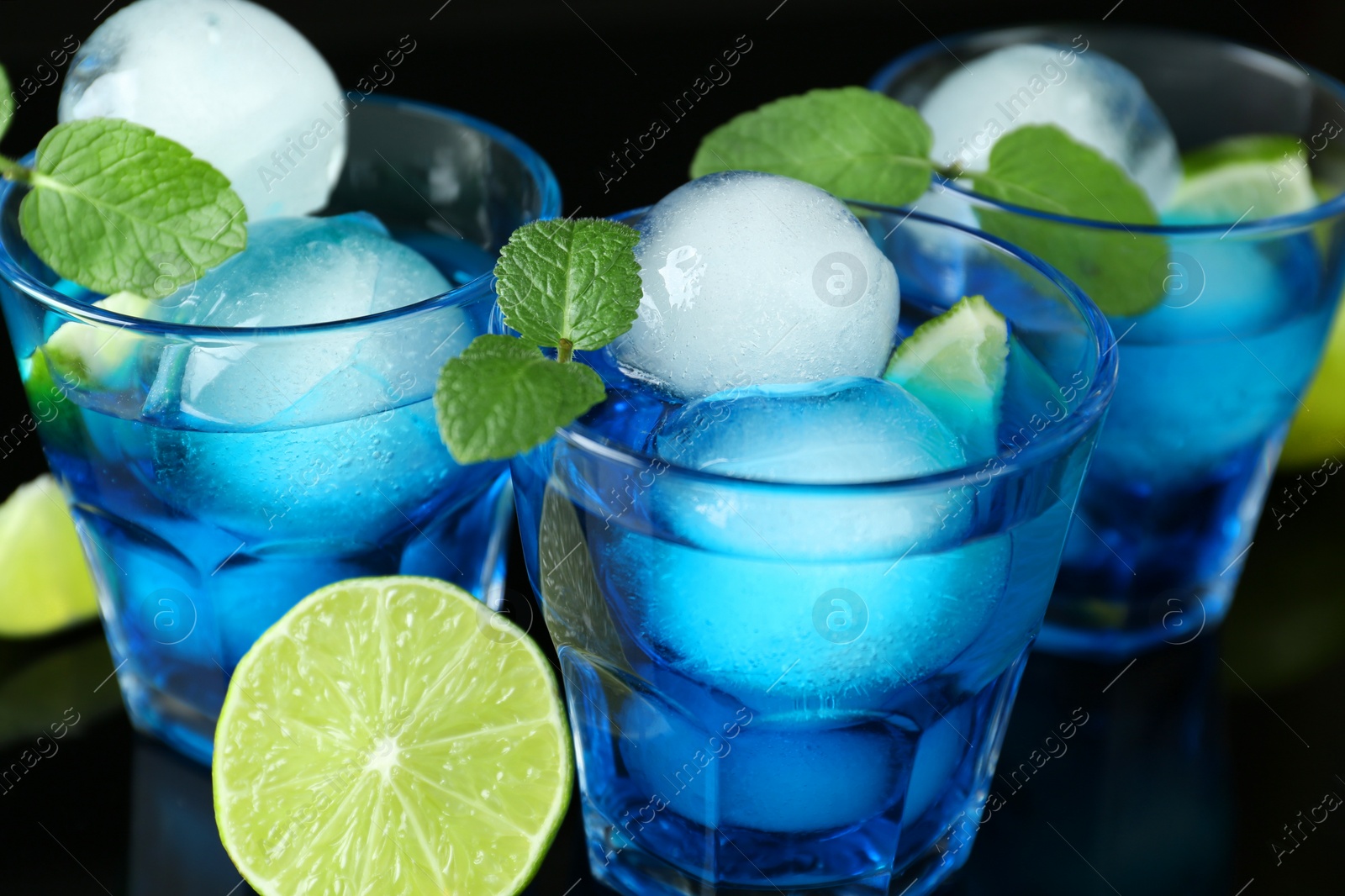 Photo of Delicious cocktails with ice balls, mint and lime on black background, closeup