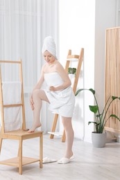 Beautiful young woman applying body cream onto leg indoors