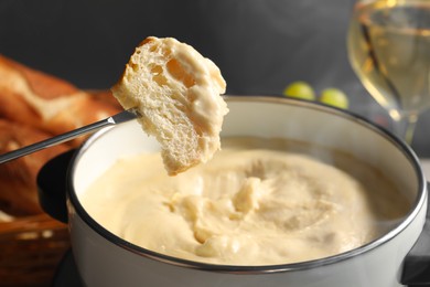 Dipping piece of bread into fondue pot with melted cheese on grey background, closeup