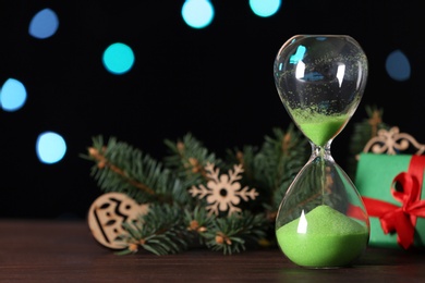 Photo of Hourglass, gift, fir tree twig and decor on wooden table against blurred lights, space for text. Christmas countdown