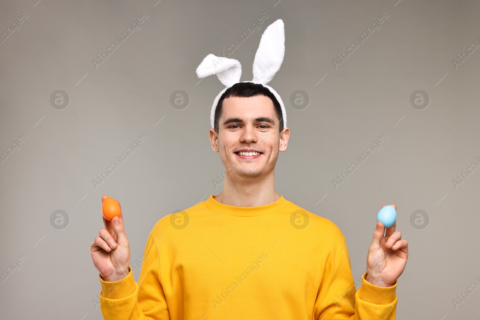 Photo of Easter celebration. Handsome young man with bunny ears holding painted eggs on grey background