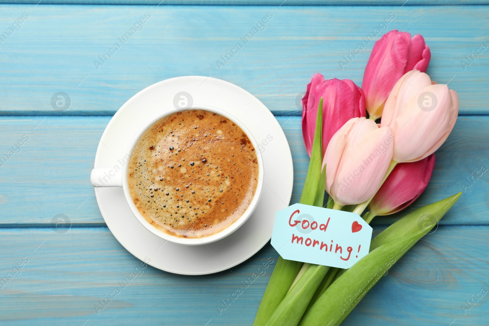 Photo of Cup of aromatic coffee, beautiful pink tulips and Good Morning note on light blue wooden table, flat lay