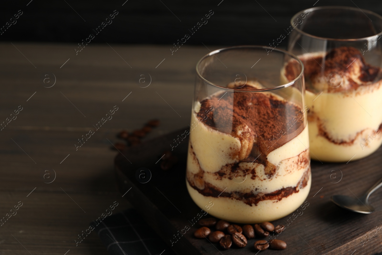 Photo of Delicious tiramisu in glasses, spoon and coffee beans on wooden table, closeup. Space for text