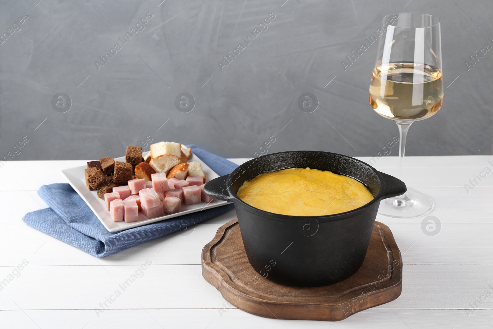 Photo of Fondue with tasty melted cheese, pieces of bread, ham and aromatic wine in glass on white wooden table