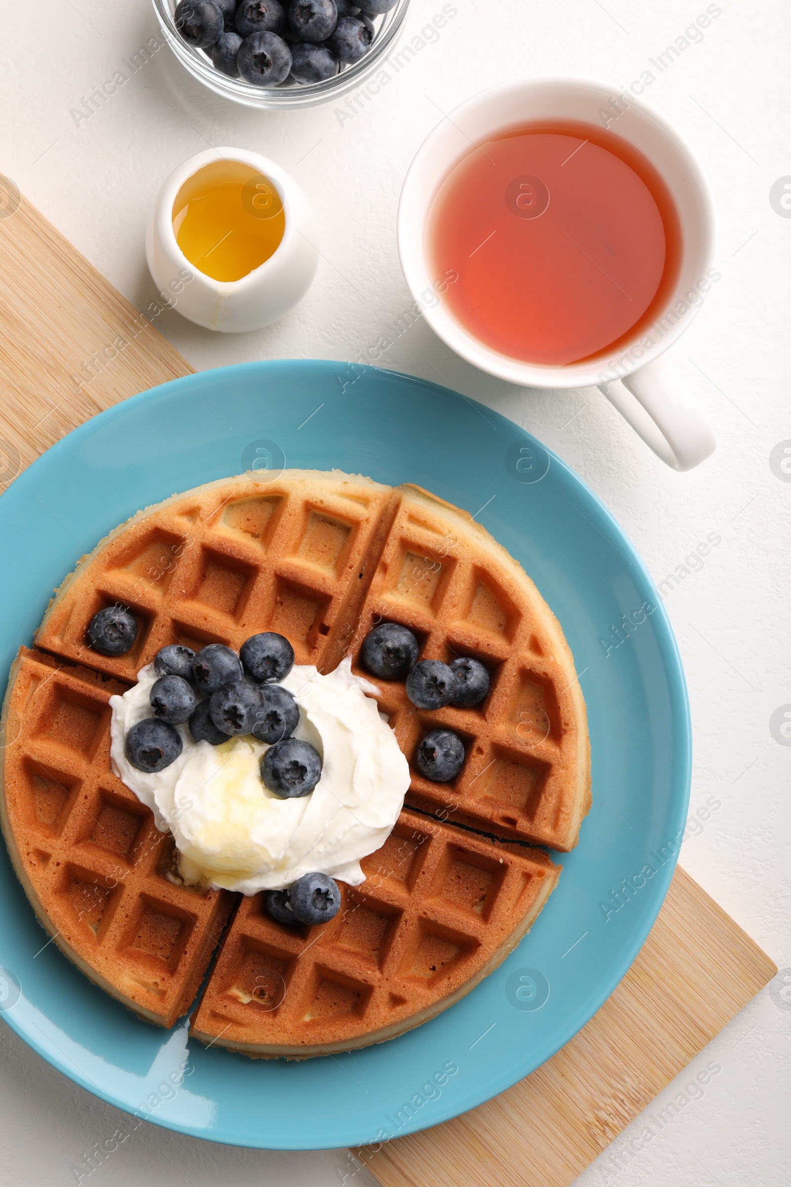 Photo of Tasty Belgian waffle with blueberries, honey and whipped cream served on white table, flat lay