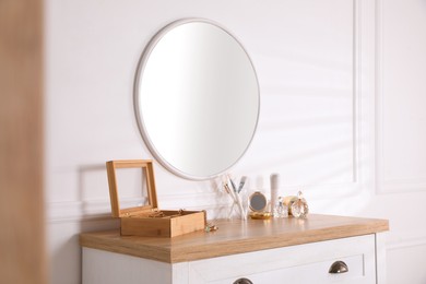 Trendy round mirror and chest of drawers with accessories near white wall indoors.