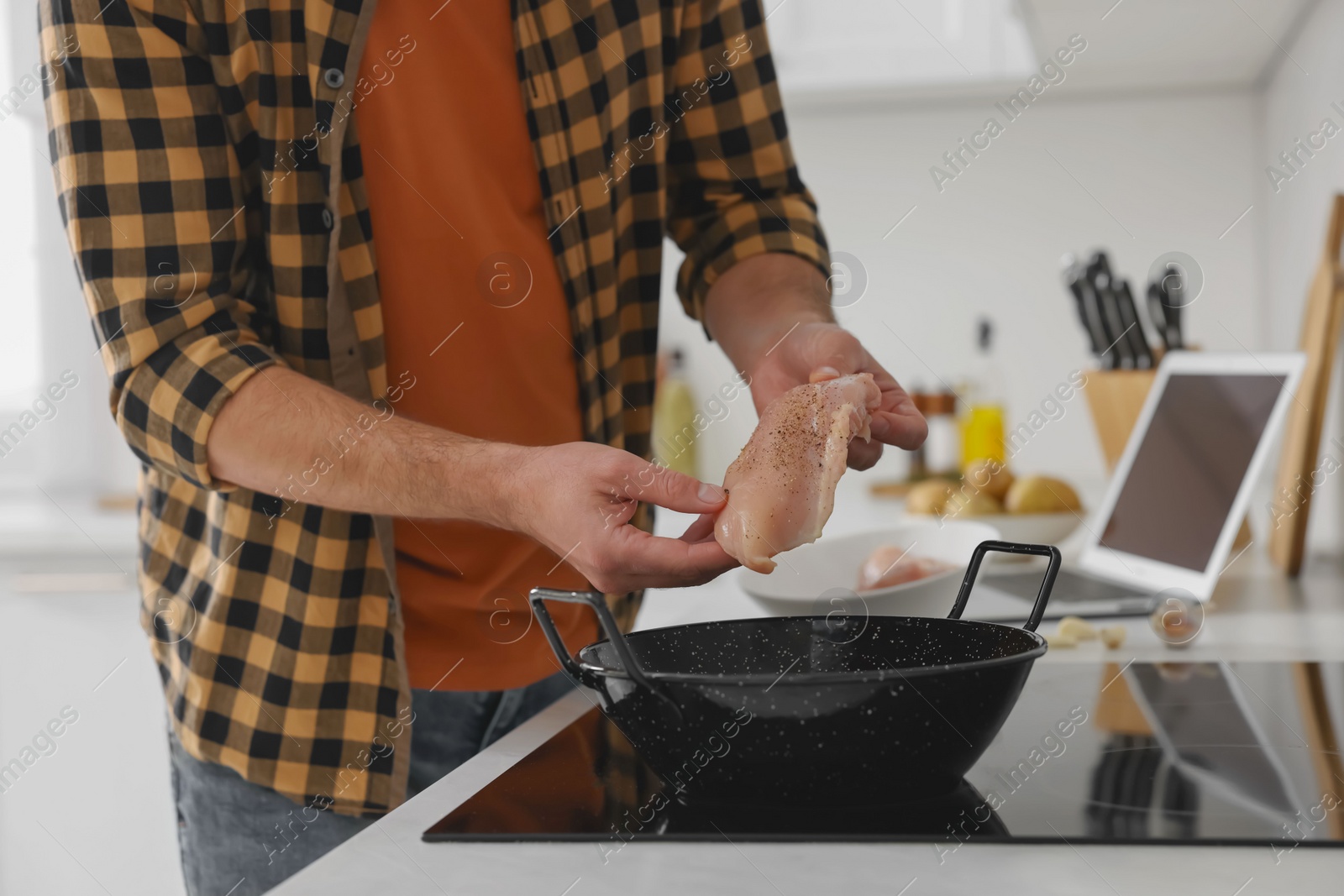 Photo of Man putting chicken fillet into frying pan while watching online cooking course via laptop in kitchen, closeup