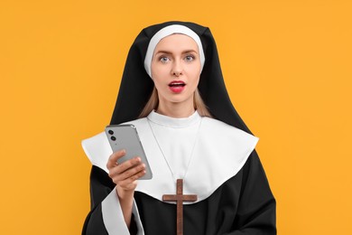 Photo of Surprised woman in nun habit with smartphone against orange background