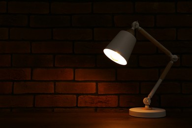 Photo of Stylish modern desk lamp on wooden table near brick wall at night, space for text
