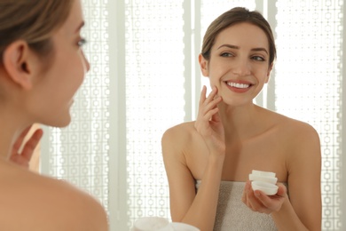 Photo of Young woman applying cream onto her face near mirror in bathroom
