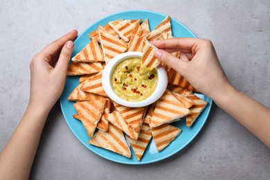 Woman dipping pita chip into hummus at grey table, top view