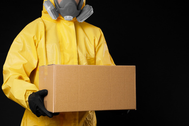 Man wearing chemical protective suit with cardboard box on black background, closeup. Prevention of virus spread