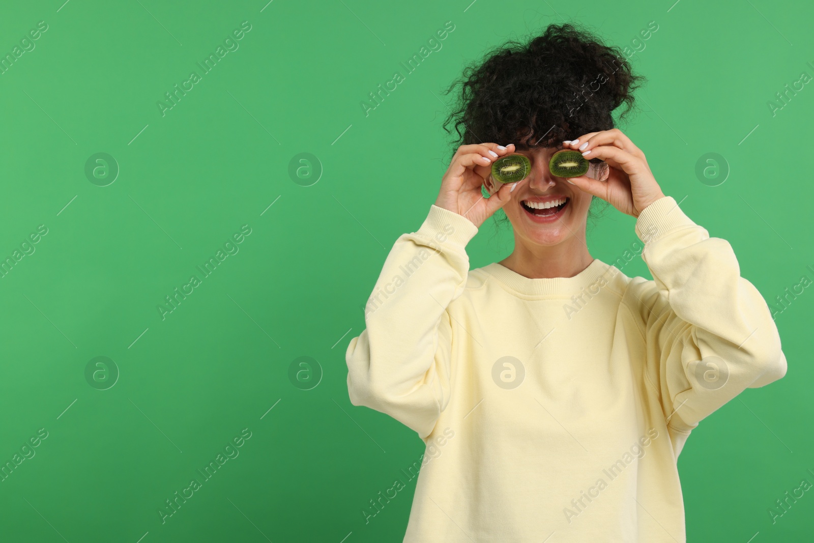 Photo of Woman covering eyes with halves of kiwi on green background, space for text