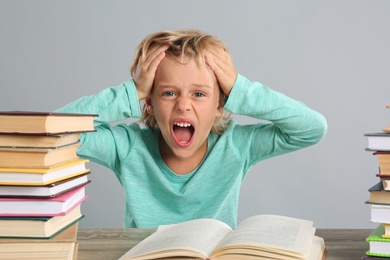 Emotional little boy doing homework at table on grey background