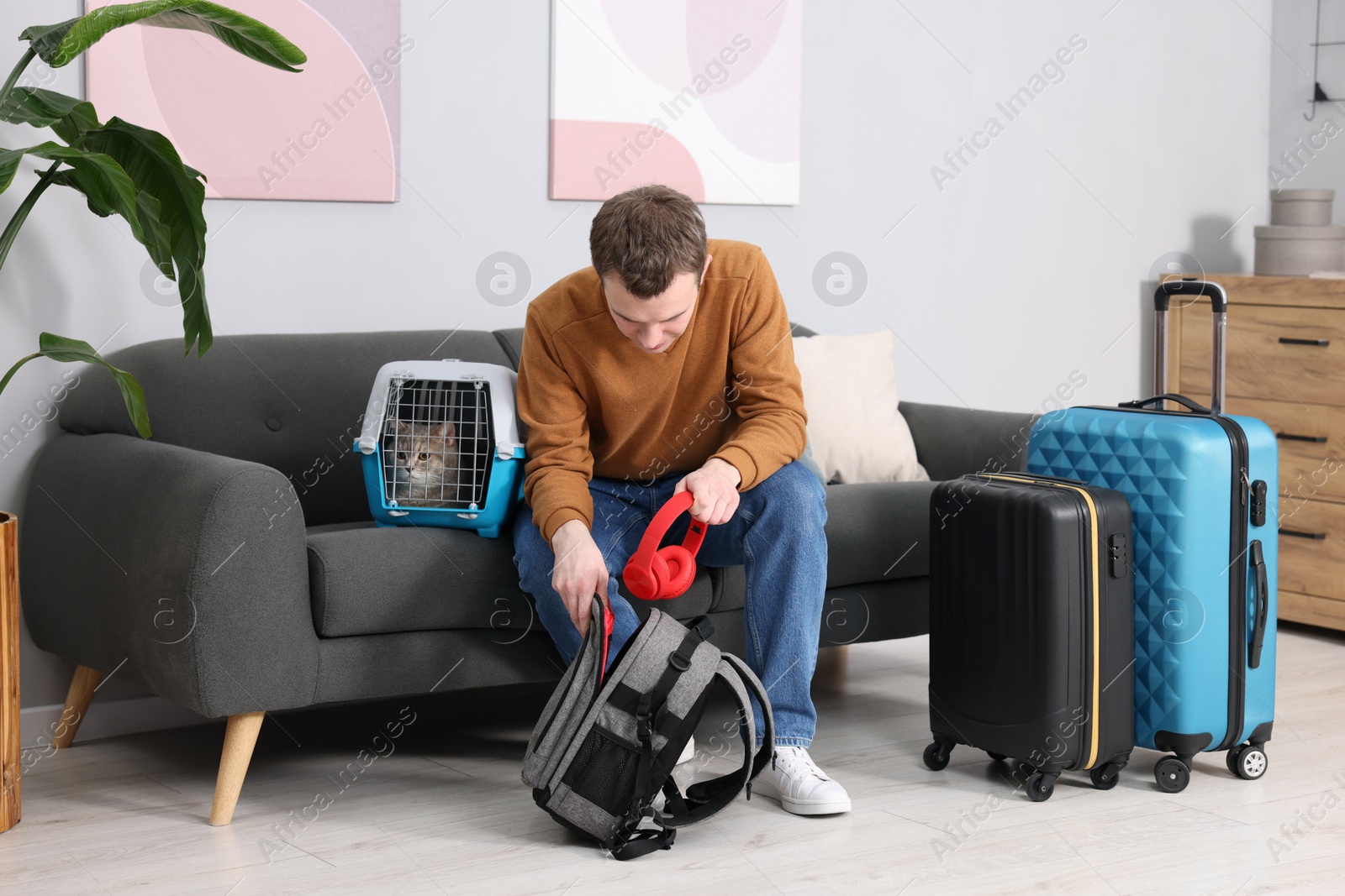 Photo of Travel with pet. Man putting headphones into backpack near carrier with cute cat at home