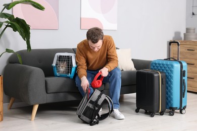 Photo of Travel with pet. Man putting headphones into backpack near carrier with cute cat at home