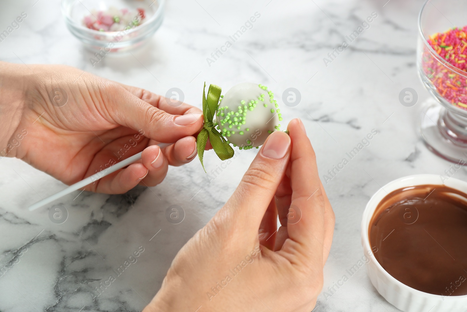 Photo of Young woman decorating tasty cake pop with green sprinkles at white marble table, closeup