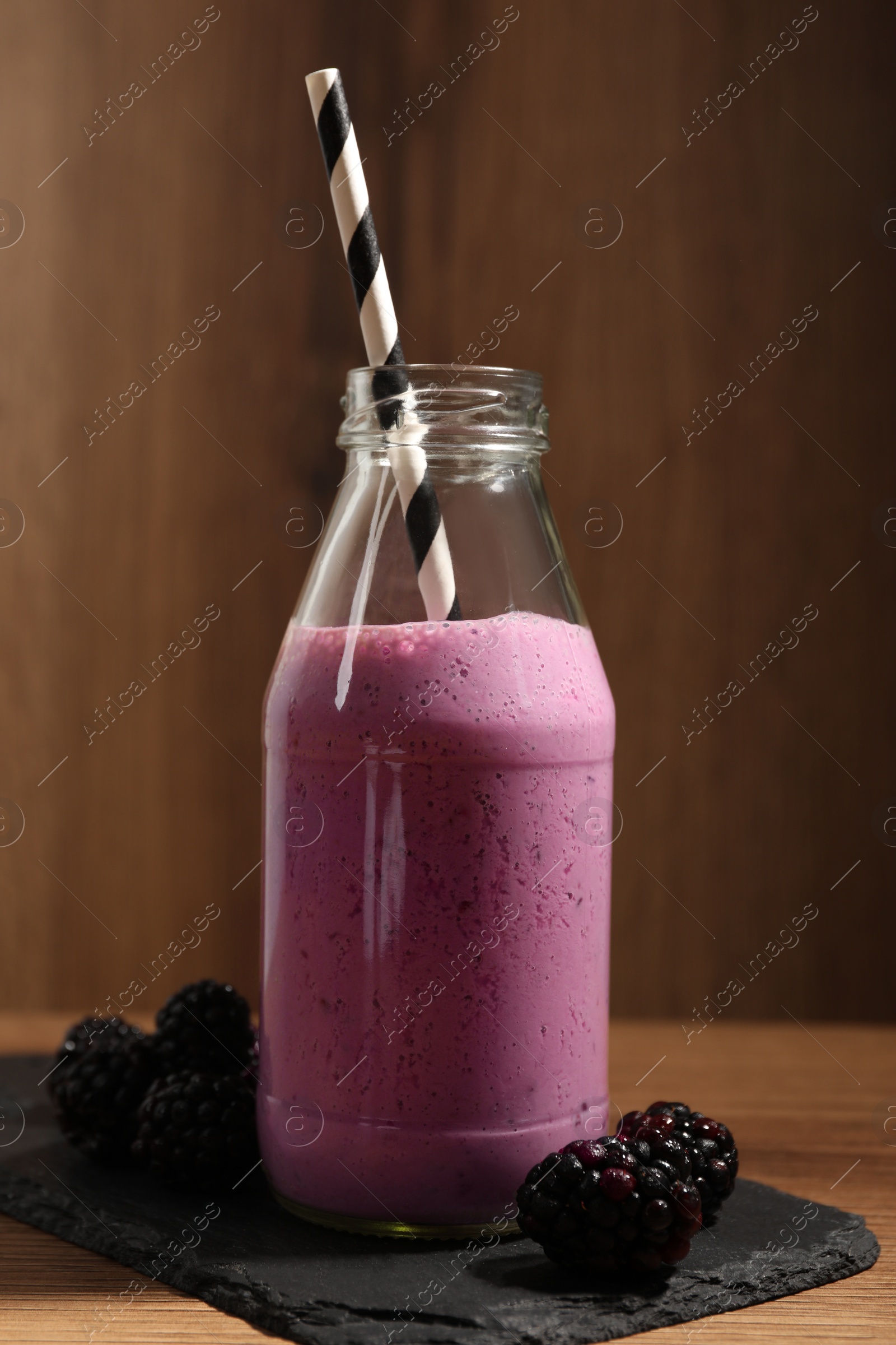 Photo of Delicious blackberry smoothie in glass bottle with straw and fresh berries on wooden table