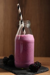 Photo of Delicious blackberry smoothie in glass bottle with straw and fresh berries on wooden table