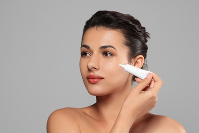Woman applying cream under eyes on grey background, space for text. Skin care