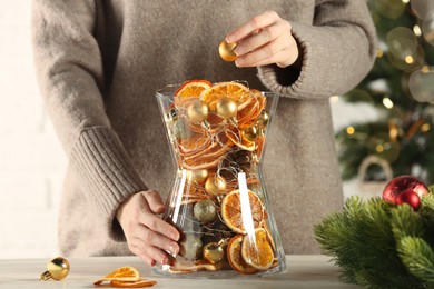 Photo of Woman putting Christmas tree ball into glass vase with dry orange slices and cones at white wooden table, closeup, Festive decor