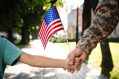 Photo of Soldier and his little son with American flag holding hands outdoors, closeup. Veterans Day in USA