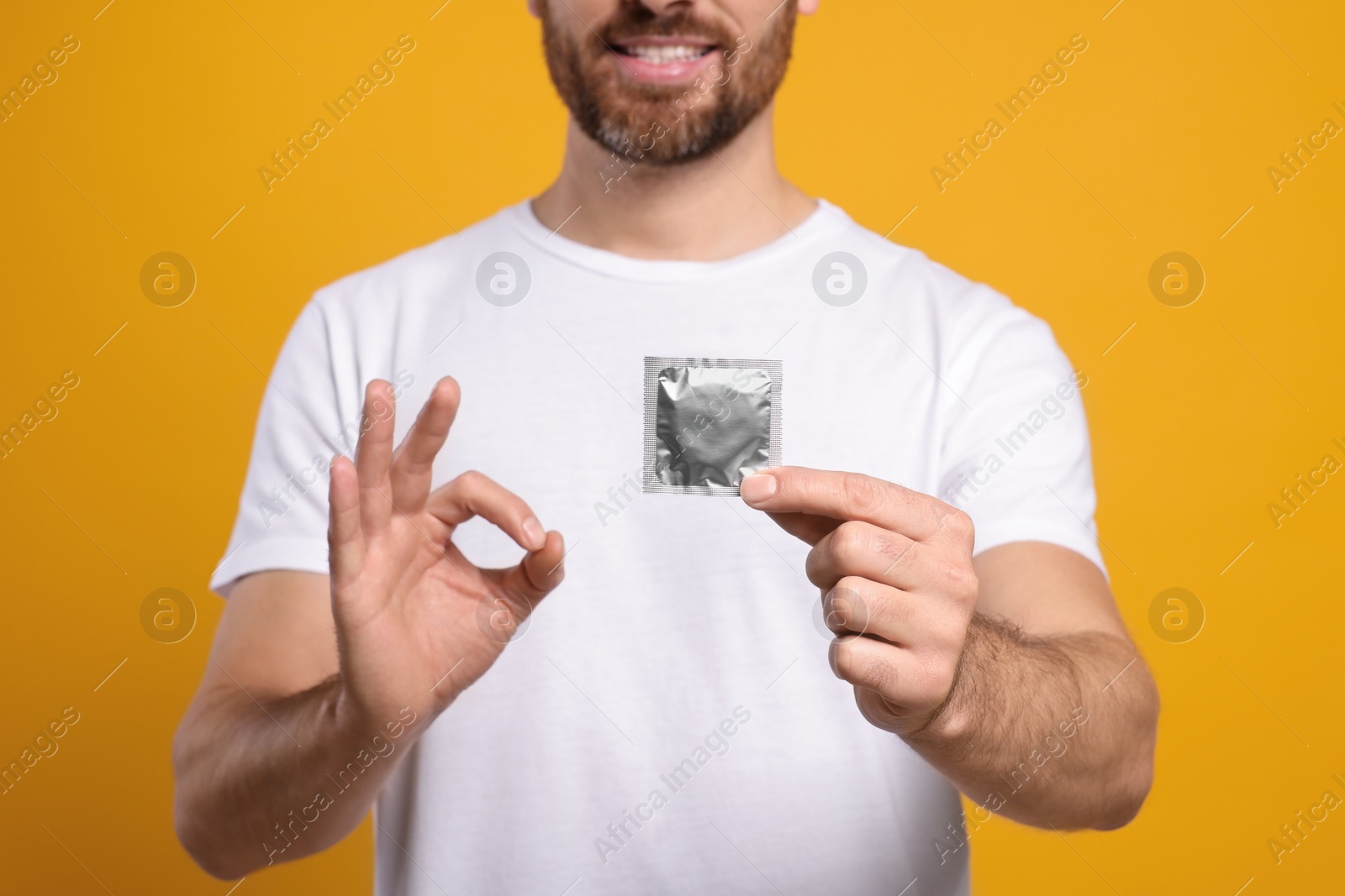 Photo of Man with condom showing ok gesture on yellow background, closeup. Safe sex