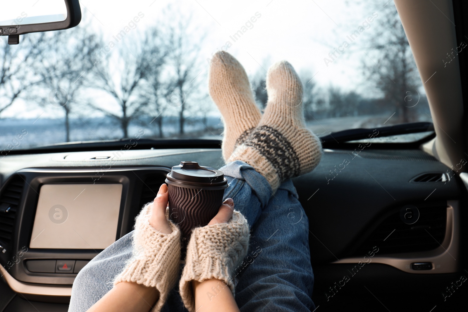 Photo of Young woman in warm socks holding her legs on car dashboard and drinking coffee. Cozy atmosphere