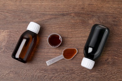 Photo of Bottles of syrup, measuring cup and dosing spoon on wooden table, flat lay. Cold medicine