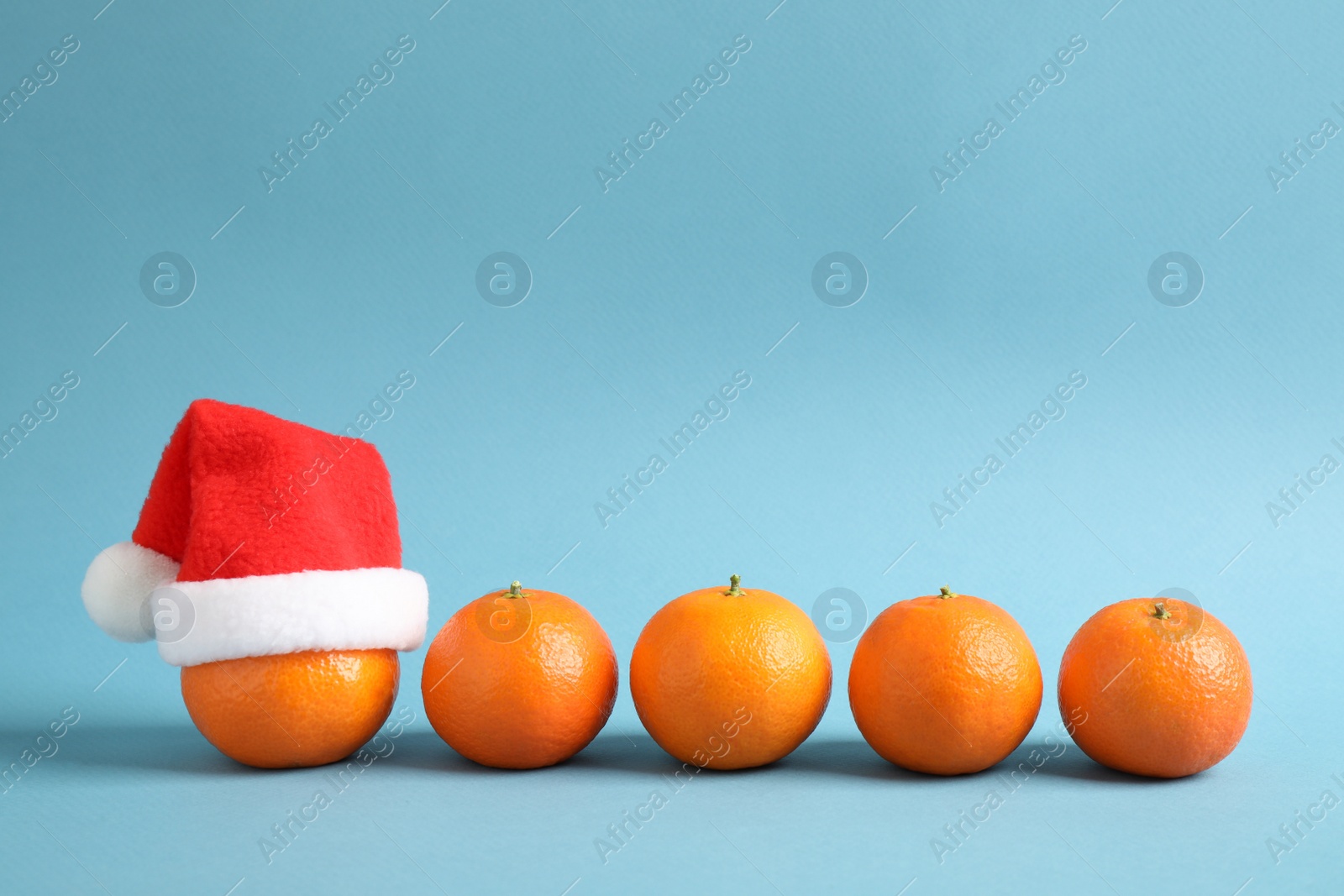 Photo of Row of fresh tangerines and one with Christmas hat against light blue background. Space for text