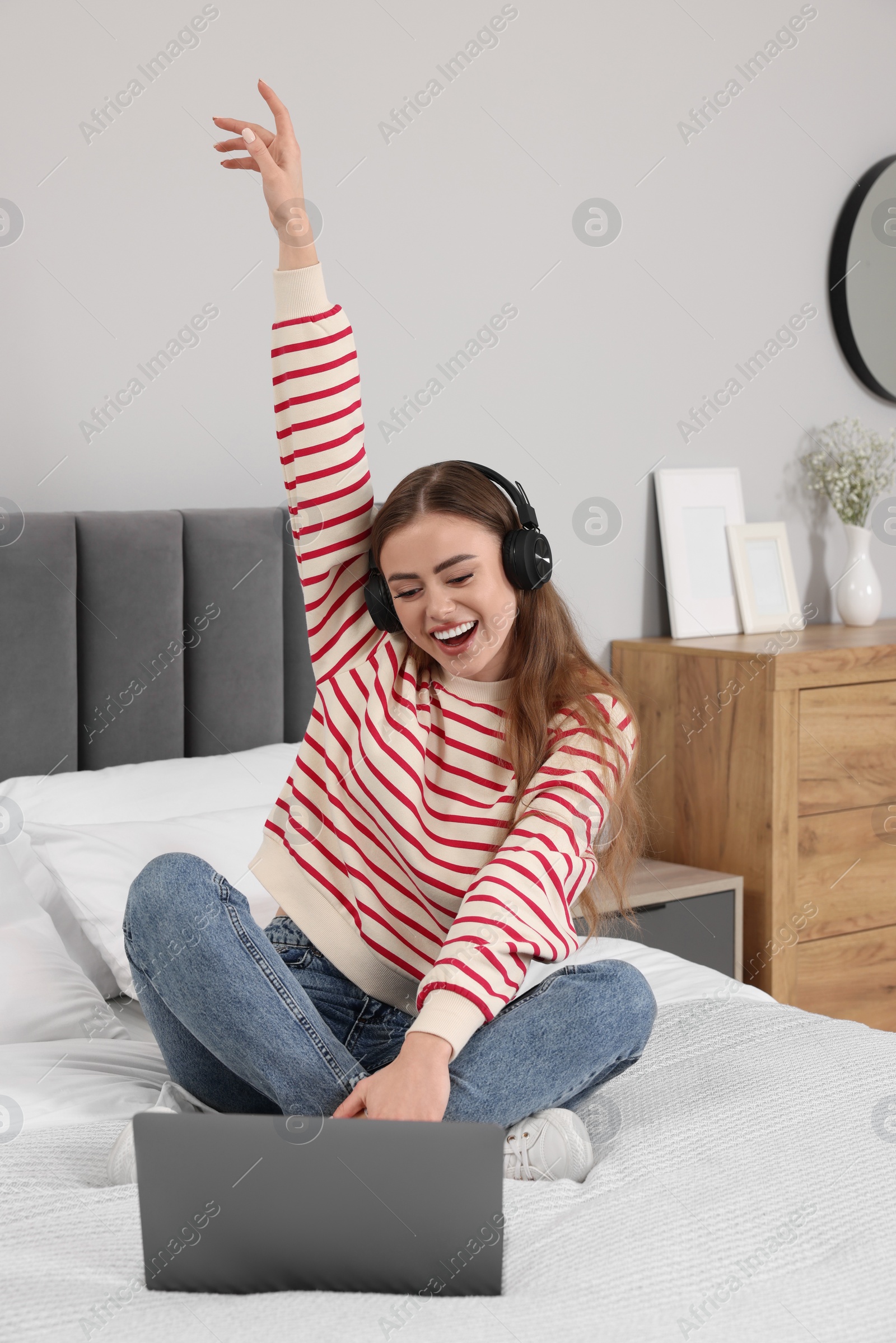 Photo of Happy woman with headphones listening to music near laptop on bed in bedroom