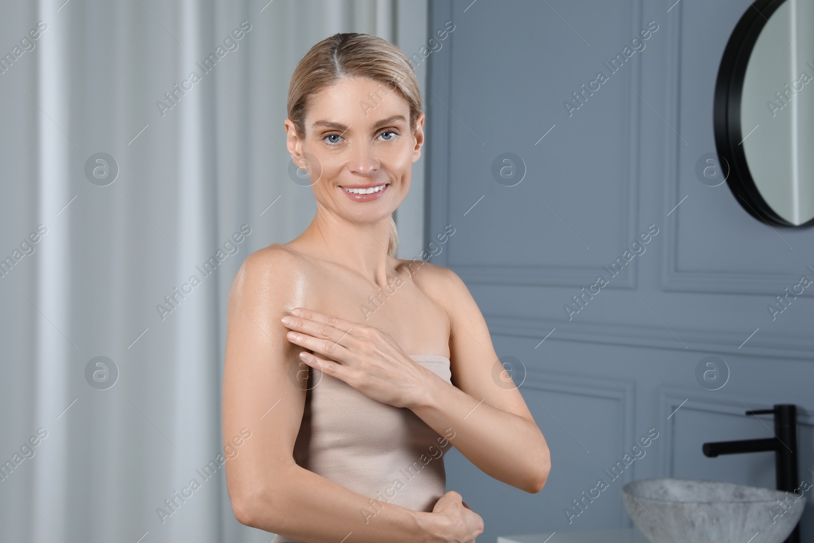 Photo of Happy woman applying body oil onto arm in bathroom